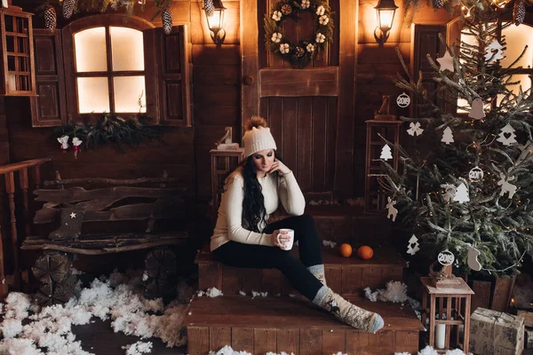 Morena em chapéu com bebida quente no alpendre de madeira sob queda de neve . — Fotografia de Stock