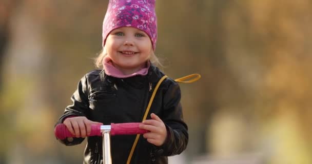 Schattig klein meisje met een scooter zwaaien op camera. — Stockvideo