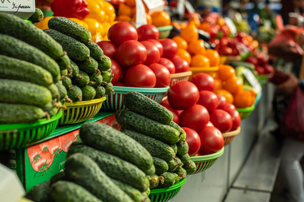 A lot of tomatoes lie on top of each other in the shape of a pyramid in the sunlight near the big green cucumbers