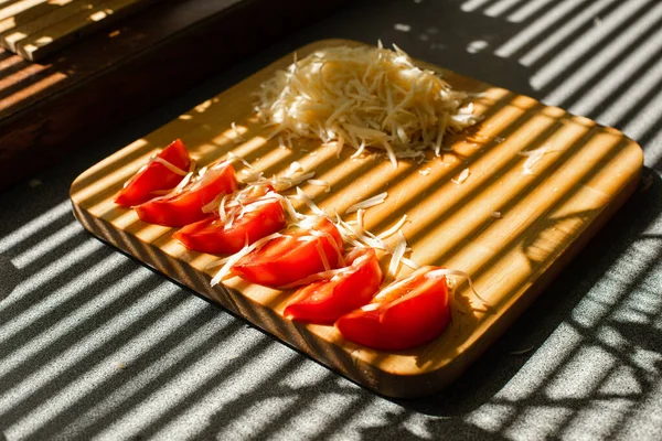 Na dřevěné desce v kuchyni leží malá hromádka strouhaného čerstvého sýra a červených rajčat. — Stock fotografie