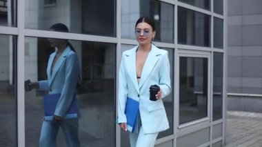 Güzel bir yüzü ve ince bir vücudu olan genç bir kadın ofisin yanında yürüyor ve kahve içiyor.