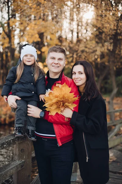 Kavkazská rodina v podzimním parku stojí na mostě proti rozmazaným žlutým stromům. — Stock fotografie