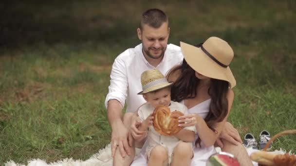 Os pais jovens dão um pão a uma criança em um piquenique no parque no verão. Instrução — Vídeo de Stock