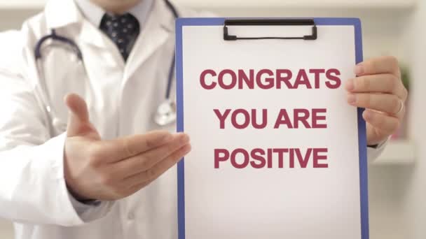 Dokter draagt een stethoscoop wijzend naar een klembord met CONGRATS U POSITIEVE titel als diagnose schot — Stockvideo
