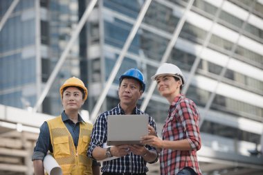 Asya mühendisler grubu dizüstü bilgisayar ve planı kağıt basılı tutarak çalışma sitesi yapı inşaat bakın. Şehir arka planda. Teknoloji, ekip çalışması kavramı