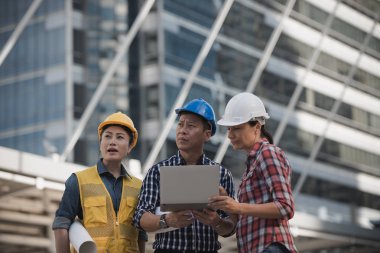 Asya mühendisler grubu dizüstü bilgisayar ve planı kağıt basılı tutarak çalışma sitesi yapı inşaat bakın. Şehir arka planda. Teknoloji, ekip çalışması kavramı
