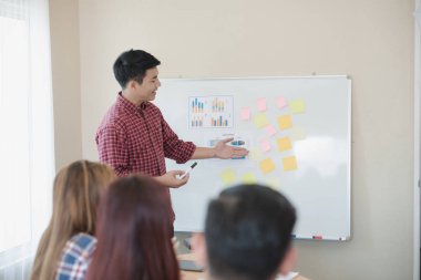 Ekose gömlek giyen yakışıklı genç iş adamı. O, ofiste kağıt beyaz tahtanın yanında sunum yapıyor, seyircilerine bakıyor ve gülümsüyor. İş toplantısı