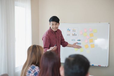 Ekose gömlek giyen yakışıklı genç iş adamı. O, ofiste kağıt beyaz tahtanın yanında sunum yapıyor, seyircilerine bakıyor ve gülümsüyor. İş toplantısı
