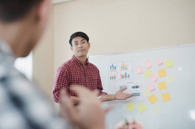 Ekose gömlek giyen yakışıklı genç iş adamı. O, ofiste kağıt beyaz tahtanın yanında sunum yapıyor, seyircilerine bakıyor ve gülümsüyor. İş toplantısı