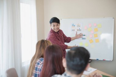 Ekose gömlek giyen yakışıklı genç iş adamı. O, ofiste kağıt beyaz tahtanın yanında sunum yapıyor, seyircilerine bakıyor ve gülümsüyor. İş toplantısı