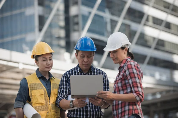 Asya mühendisler grubu dizüstü bilgisayar ve planı kağıt basılı tutarak çalışma sitesi yapı inşaat bakın. Şehir arka planda. Teknoloji, ekip çalışması kavramı