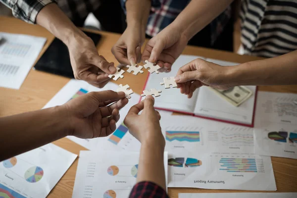 Yapboz beyazını birleştiren bir grup iş adamı. Aşağıda toplantı odasındaki ahşap masaya yerleştirilmiş grafik kağıtları var..