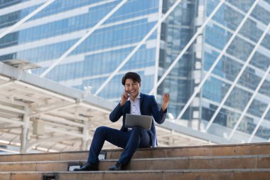 Genç Asyalı işadamı, Bangkok Tayland 'da otururken cep telefonuyla konuşmaktan ve dizüstü bilgisayarına bakmaktan memnun. İletişim kavramı