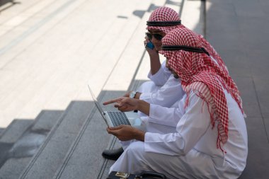 İki genç Arap işadamı, Bangkok 'un arka planında dizüstü bilgisayarla çalışırken oturup konuşuyor.