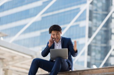 Genç Asyalı iş adamı cep telefonuyla konuşuyor ve Bangkok Tayland 'da oturan bir dizüstü bilgisayara bakıyor. İletişim kavramı