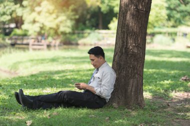 Asyalı iş adamı işten sonra parkta dinleniyor. Güneşli bir günde telefonun gölgesinin tadını çıkarıyor. İş adamı rahatlatma, dinlenme zamanı kavramı