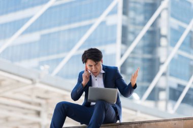 Genç Asyalı iş adamı ciddi bir yüzle cep telefonuyla konuşuyor ve Bangkok Tayland 'da dizüstü bilgisayarında çalışıyor. İletişim kavramı