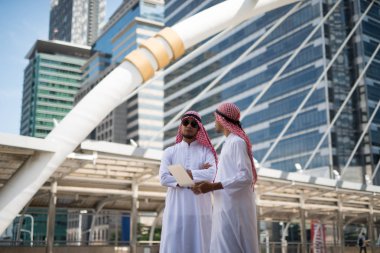 İki genç Arap işadamı, Bangkok şehrinin arka planında dizüstü bilgisayarla çalışırken konuşuyorlardı.