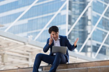 Genç Asyalı iş adamı ciddi bir yüzle cep telefonuyla konuşuyor ve Bangkok Tayland 'da dizüstü bilgisayarında çalışıyor. İletişim kavramı