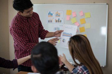 Odanın önünde dikilen sıradan ekose gömlekli genç Asyalı erkekler, konferansta başarılı bir iş yürütme konusunda meslektaşlarından geri bildirim istiyor. Takım çalışması iş toplantısı