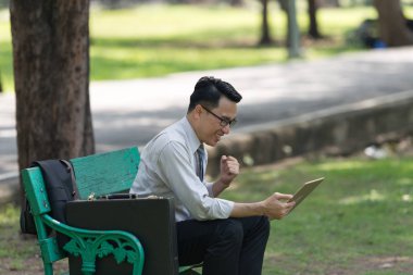 Genç işadamı parkta dijital tablet bakarak. Onların başarısı sevinç belirtileri gösteriyor. Başarılı işadamı kavramı