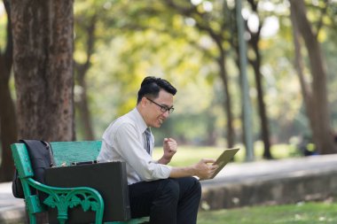 Genç işadamı parkta dijital tablet bakarak. Onların başarısı sevinç belirtileri gösteriyor. Başarılı işadamı kavramı