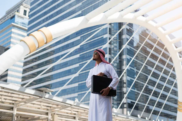 Genç Arap işadamı şehirde bir çanta tutuyor. Bangkok şehrinin arka planında, yüksek binaların olduğu iş bölgesinde,