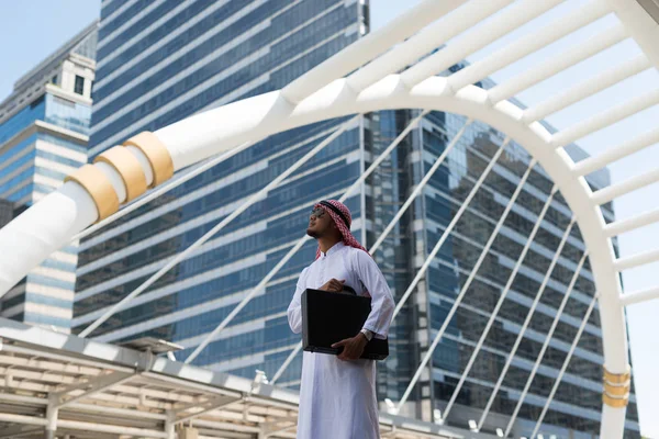 Genç Arap işadamı şehirde bir çanta tutuyor. Bangkok şehrinin arka planında, yüksek binaların olduğu iş bölgesinde,