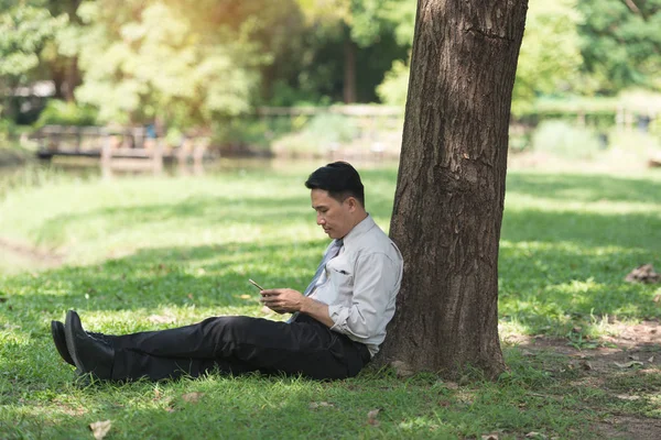 Asyalı iş adamı işten sonra parkta dinleniyor. Güneşli bir günde telefonun gölgesinin tadını çıkarıyor. İş adamı rahatlatma, dinlenme zamanı kavramı