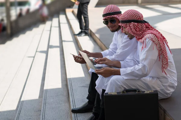 两名年轻的阿拉伯商人坐在那里聊天 一边和笔记本电脑一起工作 一边谈论邦戈曼城的背景 — 图库照片