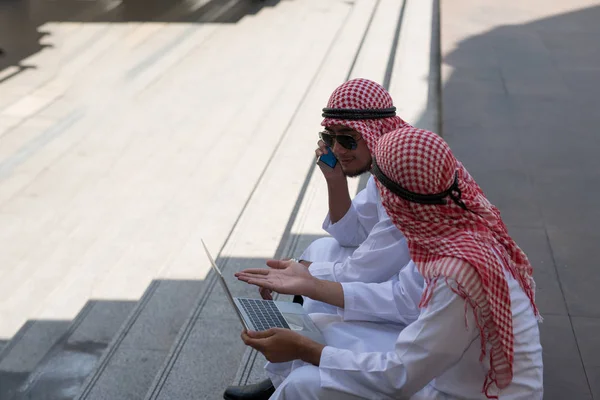 İki genç Arap işadamı, Bangkok 'un arka planında dizüstü bilgisayarla çalışırken oturup konuşuyor.