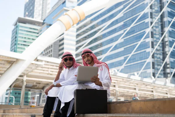 两名年轻的阿拉伯商人坐在一起聊天 一边与笔记本电脑合作 一边在大楼高楼大厦的商业区 孟加拉城的背景下工作 — 图库照片