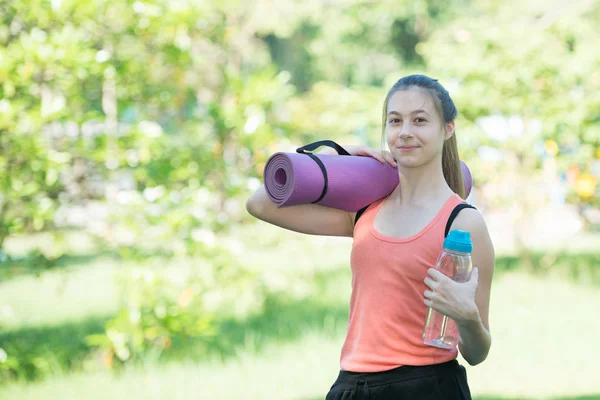 Güzel genç sporcumuz bir şişe su ve Yoga Mat parkta size gülümseme olmak tutuyor. Sağlık ve Fitness kavramı