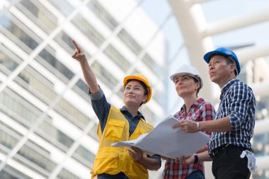 Asya mühendisleri grup planı kağıt basılı tutarak çalışma sitesi yapı inşaat bakın. Şehir arka planda. ekip çalışması kavramı