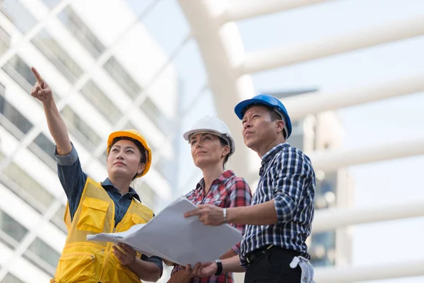 Asya mühendisleri grup planı kağıt basılı tutarak çalışma sitesi yapı inşaat bakın. Şehir arka planda. ekip çalışması kavramı