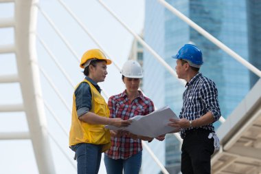 Asya mühendisleri grup planı kağıt basılı tutarak çalışma sitesi yapı inşaat bakın. Şehir arka planda. ekip çalışması kavramı