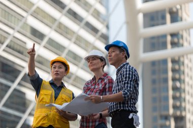 Asya mühendisleri grup planı kağıt basılı tutarak çalışma sitesi yapı inşaat bakın. Şehir arka planda. ekip çalışması kavramı