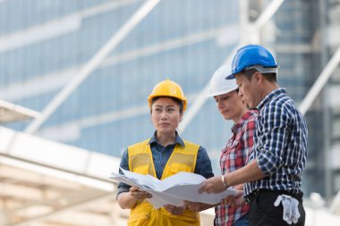 Asya mühendisleri grup planı kağıt basılı tutarak çalışma sitesi yapı inşaat bakın. Şehir arka planda. ekip çalışması kavramı