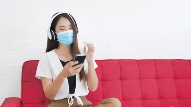 两名亚洲少女坐在沙发上 戴着医疗面罩 在家里用智能手机听音乐 新的正常 社会疏离和病毒保护概念 多利滑翔机射击 — 图库视频影像
