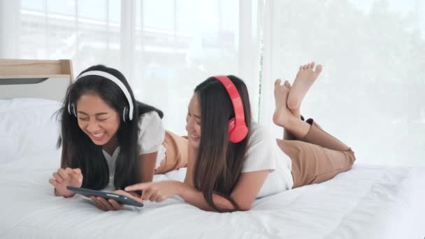两名在耳机中快乐地使用数码平板电脑的亚洲少女躺在床上听着家里的音乐 妇女放松 生活方式 青春和快乐的概念 — 图库视频影像