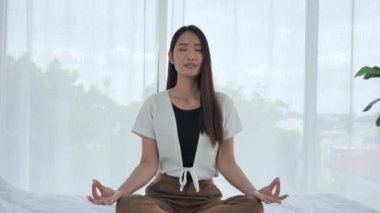 Gündelik giysiler içindeki Asyalı genç kız, bilgisayar laptopuyla yatakta meditasyon yapıyor. Evde yoga egzersizi yapan genç bir kadın işten dolayı stres atmak için. Dolly kaydırma atışı.