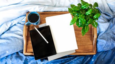 Mavi yatakta ahşap tepsi. Evde çalış. Tepside çiçekli bir fincan çay. Tablet ve telefon yatağın üstünde. Güzel modern neoklasik iç mimari.