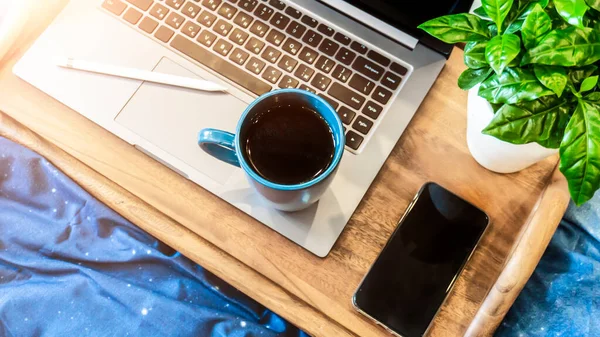 Wooden tray on a blue bed. Work at home. Cup of tea on a tray with a flower. Tablet and phone on the bed. Beautiful modern neoclassical interior.