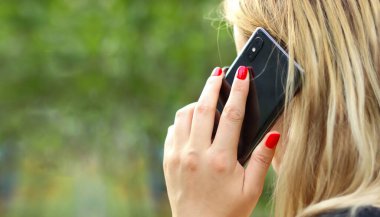 Sarışın kız telefonda konuşuyor, kırmızı manikür, uzun saç, siyah telefon. Yakın plan, bulanık yeşil arka plan. Yaşam tarzı.