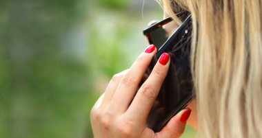 Sarışın kız telefonda konuşuyor, kırmızı manikür, uzun saç, siyah telefon. Yakın plan, bulanık yeşil arka plan. Yaşam tarzı.