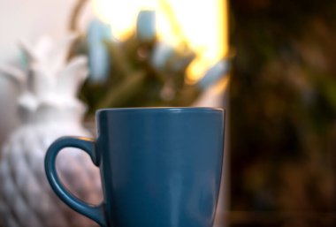 Akşam kahvesi. Mavi bardakta kahve, açık kitap ve cep telefonu, akşam ışığı, gün batımı, alacakaranlık. Atmosferik, bahçede sıcak bir akşam, yeşil yapraklar, sohbet. Sıcak ışık, bulanık bokeh.