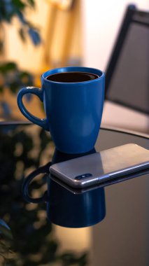 Akşam kahvesi. Mavi bardakta kahve, açık kitap ve cep telefonu, akşam ışığı, gün batımı, alacakaranlık. Atmosferik, bahçede sıcak bir akşam, yeşil yapraklar, sohbet. Sıcak ışık, bulanık bokeh.