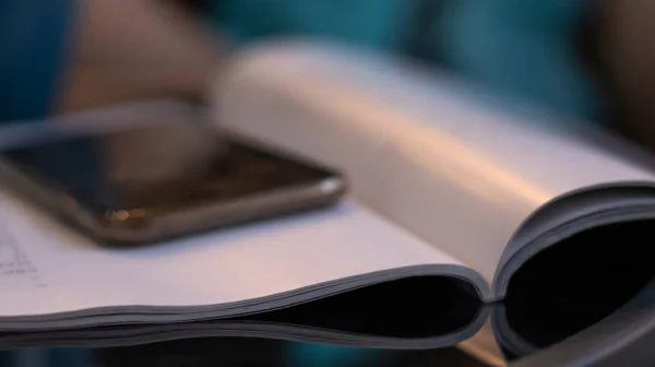 Open book and phone on a glass black table. Blurred boke background, close-up. Dark evening background with sunset highlights.