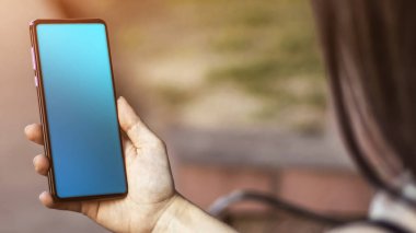 Esmer bir kız parkta oturuyor, elinde bir cep telefonu var. Güneş ışığı, bulanık arkaplan. Yaşam biçimi.