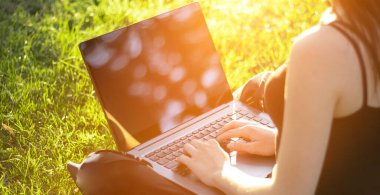 Dizüstü bilgisayarı olan bir kız çimlere oturur, parktaki çimlere oturur. Çalışmak, temiz havada çalışmak. Bokeh bulanıklığı, güneş ışığı.